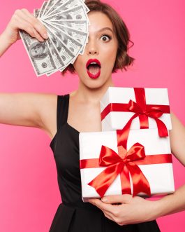 Portrait of a cheerful girl dressed in black dress holding stack of gift boxes and bunch of money banknotes isolated over pink background