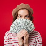 Portrait of a young woman wearing beret holding bunch of money banknotes at her face isolated over pink background