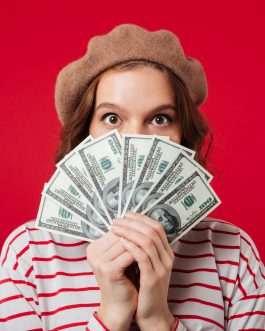 Portrait of a young woman wearing beret holding bunch of money banknotes at her face isolated over pink background